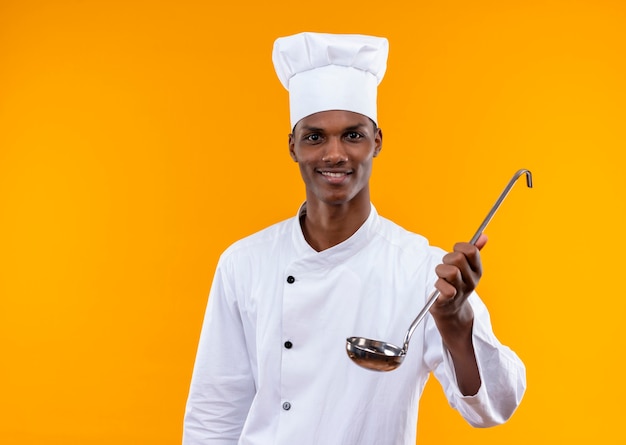 Il giovane cuoco afroamericano sorridente in uniforme del cuoco unico tiene il mestolo isolato sulla parete arancione