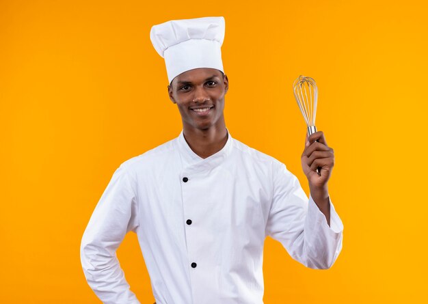 Il giovane cuoco afroamericano sorridente in uniforme del cuoco unico tiene la frusta e mette la mano sulla vita isolata sulla parete arancio