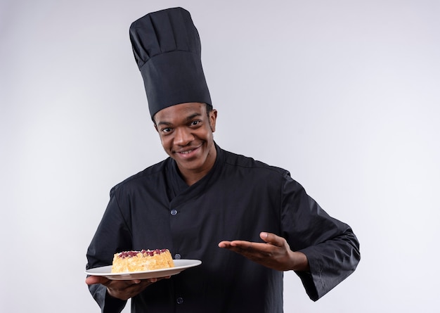 Il giovane cuoco afroamericano sorridente in uniforme del cuoco unico tiene la torta sul piatto e sui punti con la mano isolata sulla parete bianca