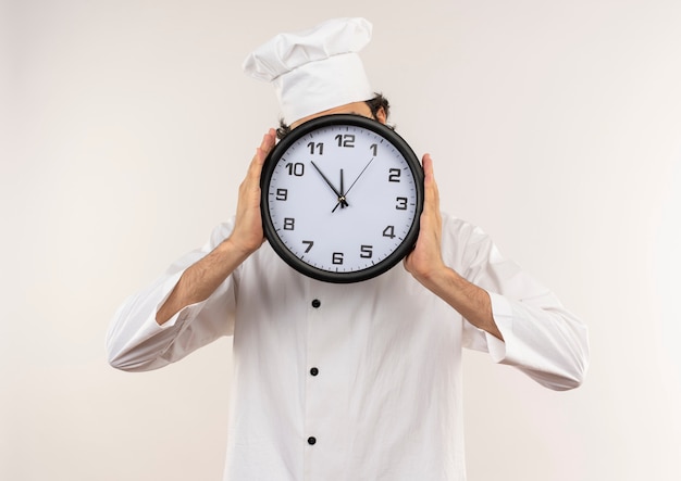 Il giovane cuoco maschio che indossa l'uniforme e gli occhiali del cuoco unico ha coperto il fronte con l'orologio di parete isolato sulla parete bianca