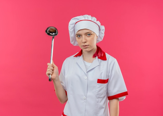 Il giovane cuoco unico femminile biondo sorpreso in uniforme del cuoco unico tiene il mestolo e sembra isolato sulla parete rosa