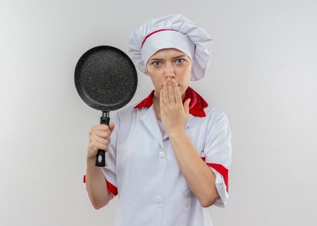 Il giovane cuoco unico femminile biondo sorpreso in uniforme del cuoco unico tiene la padella e mette la mano sulla bocca isolata sulla parete bianca