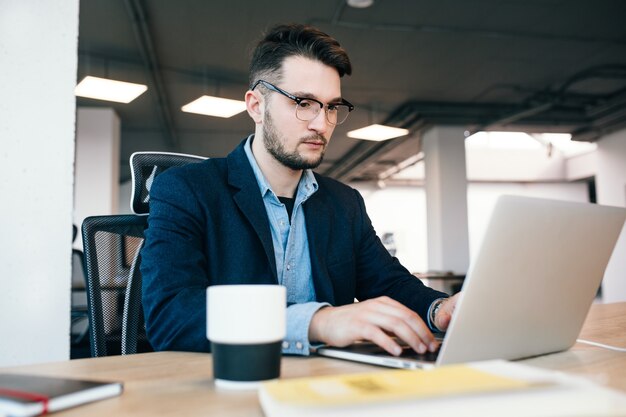 Il giovane dai capelli scuri sta lavorando al tavolo in ufficio. Indossa una camicia blu con una giacca nera. Sta scrivendo seriamente sul laptop.