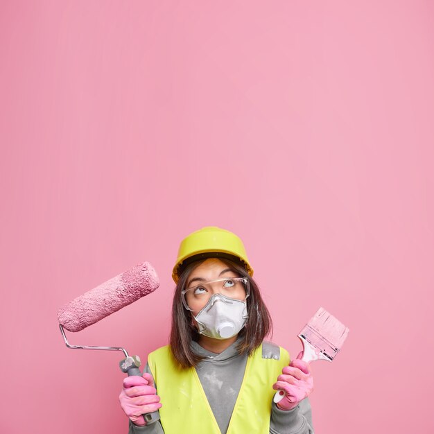Il giovane decoratore femminile asiatico professionista esitante tiene gli strumenti di pittura pensa alla ristrutturazione della casa focalizzata sopra indossa il respiratore del casco protettivo e gli occhiali di sicurezza isolati sul muro rosa
