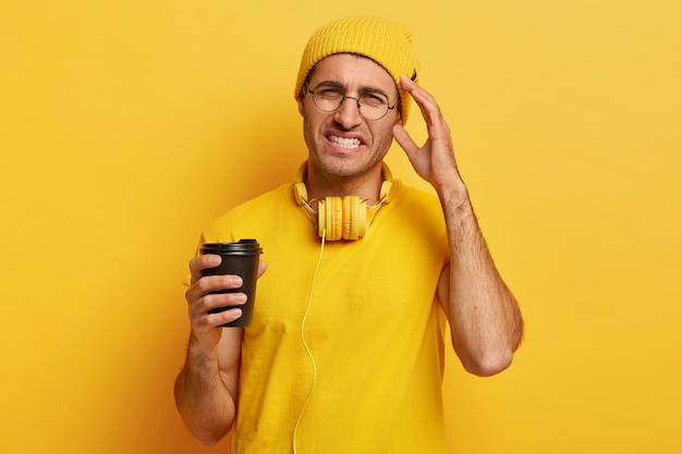 Il giovane disperato ha mal di testa, si sente oberato di lavoro e tocca la tempia, stringe i denti, indossa abiti gialli vivaci, tiene una tazza di caffè da asporto, indossa occhiali rotondi. Concetto di sentimenti negativi