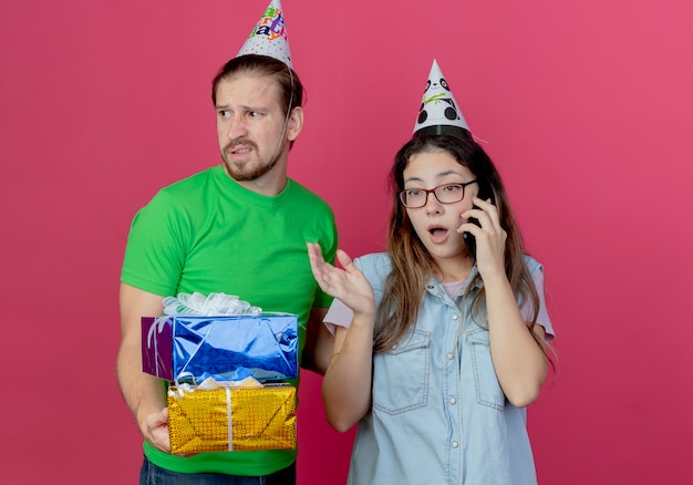 Il giovane dispiaciuto che indossa il cappello del partito tiene le scatole regalo e guarda al lato in piedi con la ragazza sorpresa che indossa il cappello del partito e parla al telefono isolato sul muro rosa