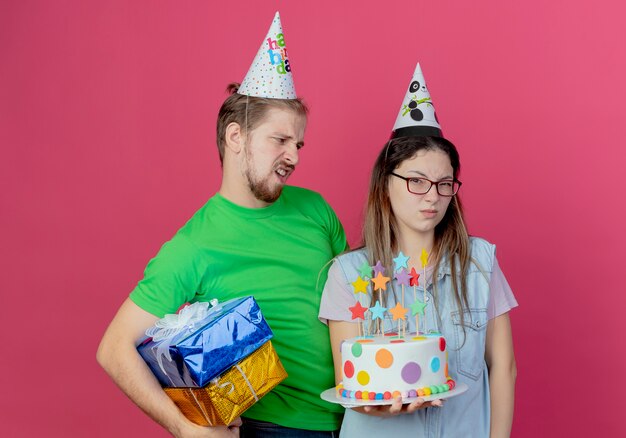 Il giovane dispiaciuto che indossa il cappello del partito tiene le scatole regalo guardando la ragazza infastidita che indossa il cappello del partito e tenendo la torta di compleanno isolata sul muro rosa