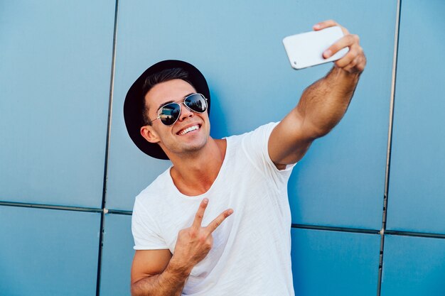 Il giovane divertente in occhiali da sole e cappello nero prende un selfie, mostrando un segno di pace