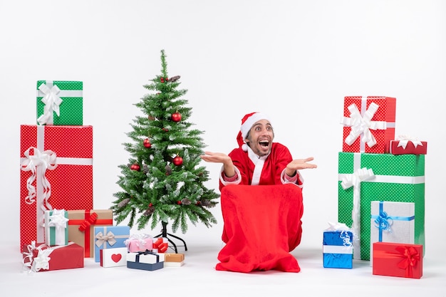 Il giovane emozionante emotivo vestito da Babbo Natale con i regali e l'albero di Natale decorato reagisce felicemente su fondo bianco