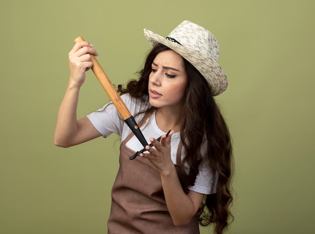 Il giovane giardiniere femminile all'oscuro in cappello da giardinaggio d'uso uniforme tiene ed esamina il rastrello isolato sulla parete verde oliva