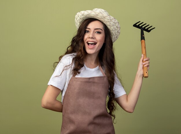 Il giovane giardiniere femminile allegro in cappello da giardinaggio d'uso uniforme tiene il rastrello isolato sulla parete verde oliva