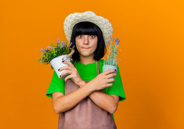 Il giovane giardiniere femminile ansioso in cappello da giardinaggio d'uso uniforme tiene i vasi di fiori che attraversano le mani