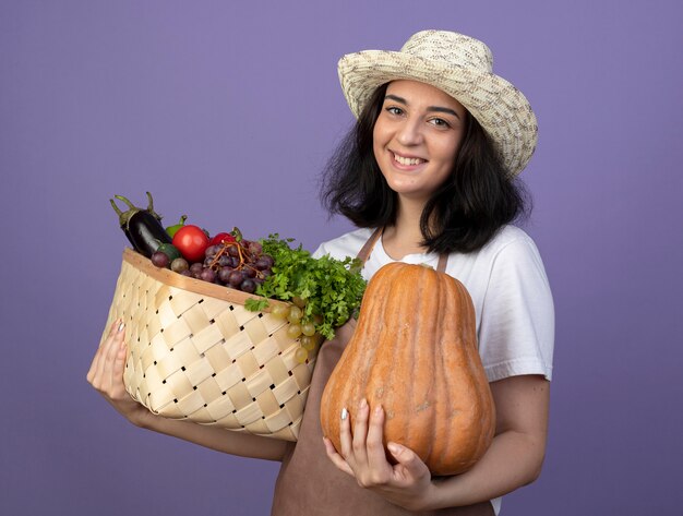 Il giovane giardiniere femminile castana sorridente in cappello da giardinaggio d'uso uniforme tiene il canestro e la zucca della verdura isolati sulla parete viola con lo spazio della copia