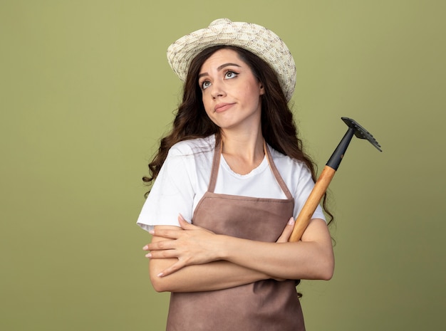 Il giovane giardiniere femminile deluso in cappello da giardinaggio da portare uniforme si leva in piedi con le braccia incrociate che tengono il rastrello isolato sulla parete verde oliva