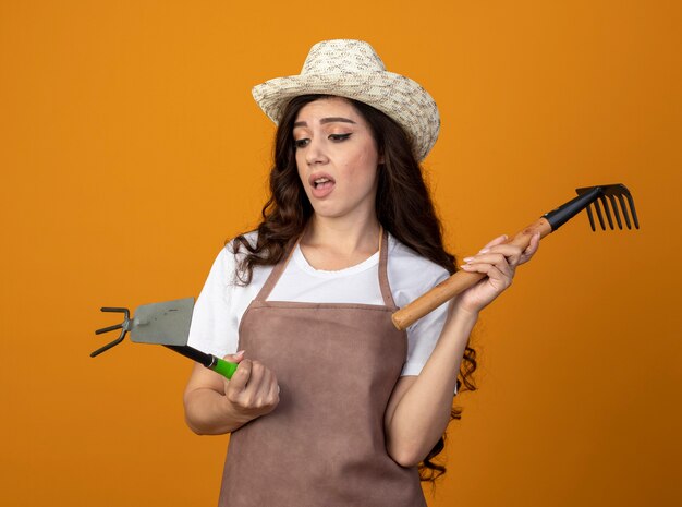 Il giovane giardiniere femminile deluso in uniforme che indossa il cappello da giardinaggio tiene il rastrello e guarda il rastrello della zappa isolato sulla parete arancione con lo spazio della copia