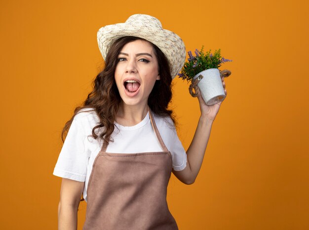 Il giovane giardiniere femminile infastidito in cappello da giardinaggio d'uso uniforme tiene il vaso di fiori isolato sulla parete arancio con lo spazio della copia
