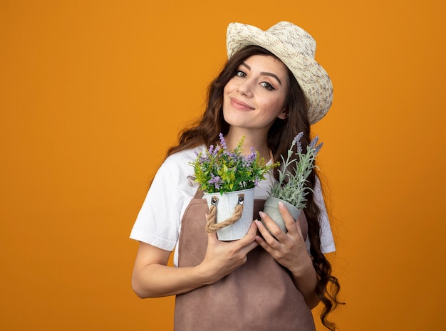 Il giovane giardiniere femminile soddisfatto in cappello da giardinaggio da portare uniforme tiene i vasi da fiori isolati sulla parete arancione con lo spazio della copia