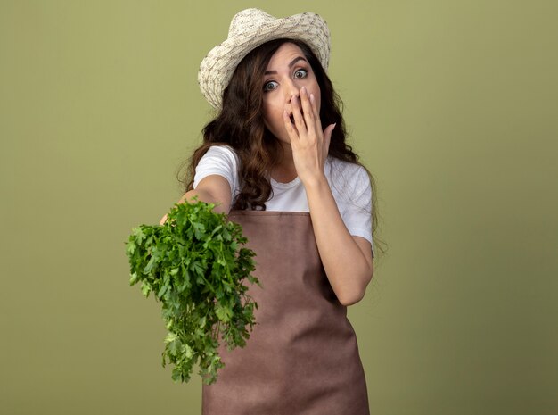 Il giovane giardiniere femminile sorpreso in cappello da giardinaggio d'uso uniforme mette la mano sulla bocca e tiene il coriandolo isolato sulla parete verde oliva