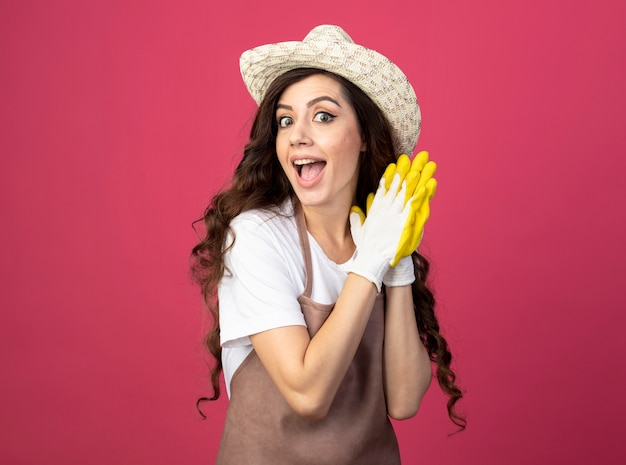 Il giovane giardiniere femminile sorpreso in cappello e guanti da giardinaggio d'uso uniformi tiene le mani insieme isolate sulla parete rosa con lo spazio della copia