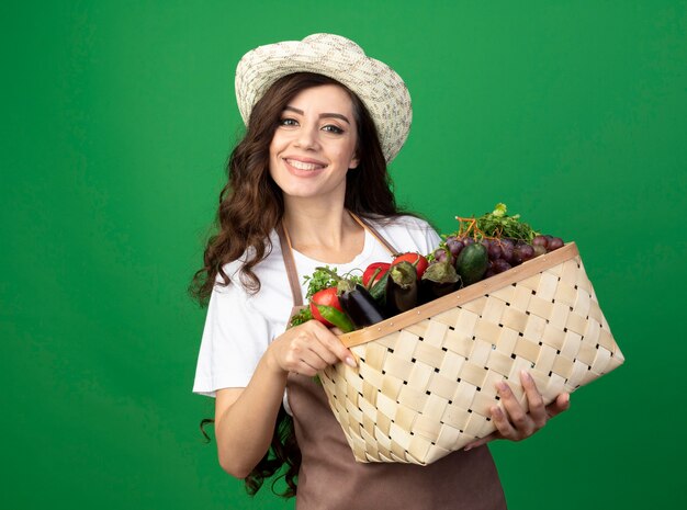 Il giovane giardiniere femminile sorridente in cappello da giardinaggio d'uso uniforme tiene il cestino della verdura isolato sulla parete verde