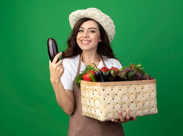 Il giovane giardiniere femminile sorridente in cappello da giardinaggio d'uso uniforme tiene il cestino e la melanzana della verdura isolati sulla parete verde