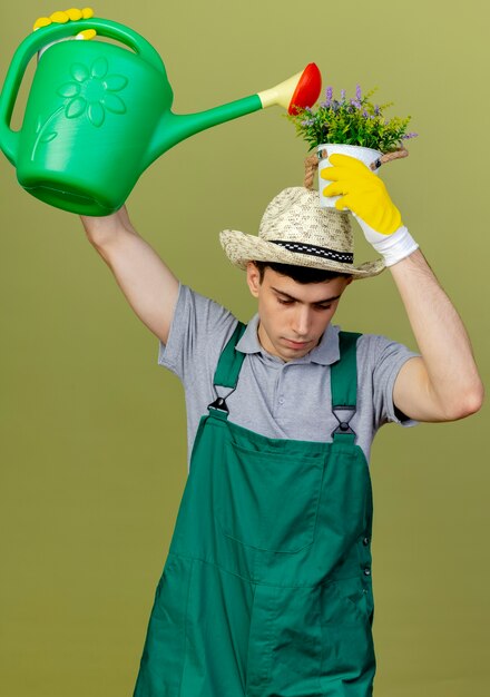 Il giovane giardiniere maschio sicuro che indossa il cappello e i guanti di giardinaggio tiene il vaso di fiori