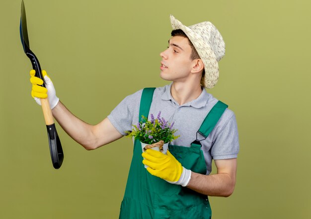 Il giovane giardiniere maschio sicuro che indossa il cappello e i guanti di giardinaggio tiene il vaso di fiori