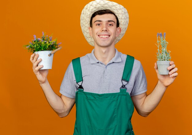 Il giovane giardiniere maschio sorridente che porta il cappello di giardinaggio tiene i fiori in vasi da fiori