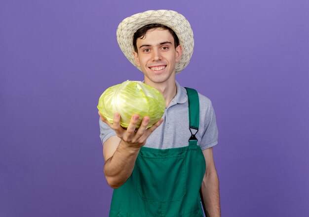 Il giovane giardiniere maschio sorridente che porta il cappello di giardinaggio tiene il cavolo