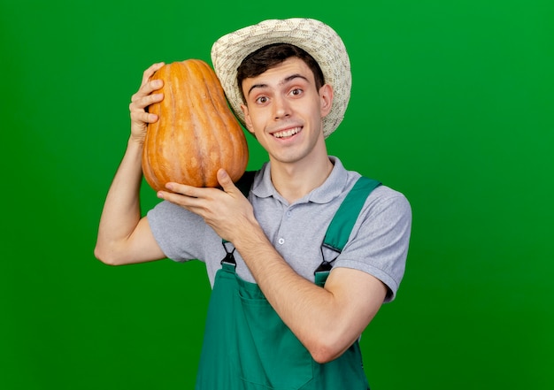 Il giovane giardiniere maschio sorridente che porta il cappello di giardinaggio tiene la zucca