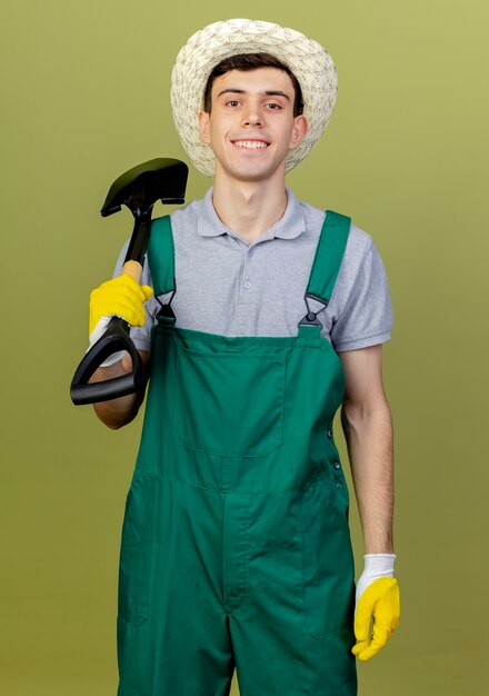 Il giovane giardiniere maschio sorridente sicuro che porta il cappello di giardinaggio tiene la vanga sulla spalla