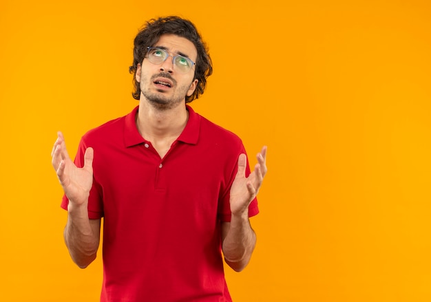 Il giovane infastidito in camicia rossa con vetri ottici tiene le mani in alto isolato sulla parete arancione