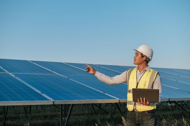 Il giovane ispettore asiatico ingegnere uomo usa il computer portatile che lavora presso la fattoria solare Supervisore tecnico maschio in casco bianco Controllo del funzionamento del sole e del pannello solare fotovoltaico nello spazio di copia della stazione