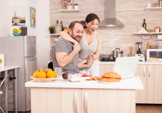 Il giovane libero professionista legge buone notizie mentre fa colazione e lavora al computer portatile in cucina. Imprenditore euforico felicissimo di successo a casa al mattino, vincitore e trionfo degli affari