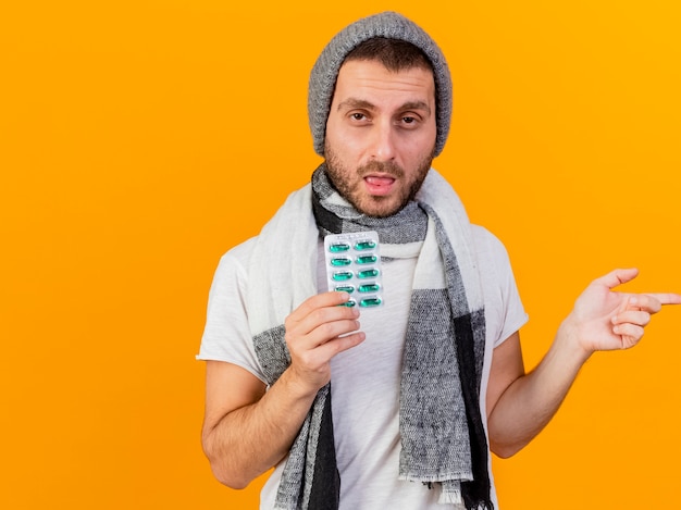 Il giovane malato dispiaciuto che indossa il cappello e la sciarpa di inverno che tengono le pillole ed i punti sul lato isolato su fondo giallo con lo spazio della copia