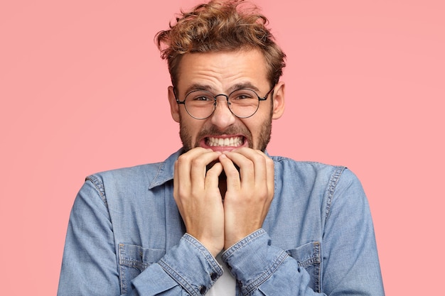 Il giovane maschio caucasico infastidito stringe i denti e guarda con espressione dispiaciuta, si morde le unghie delle dita, guarda disperatamente, prova avversione, indossa una camicia di jeans, sta contro il muro rosa. Oh no!