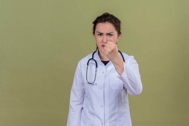 Il giovane medico arrabbiato che porta lo stetoscopio d'uso dell'abito medico mostra il pugno sulla parete verde