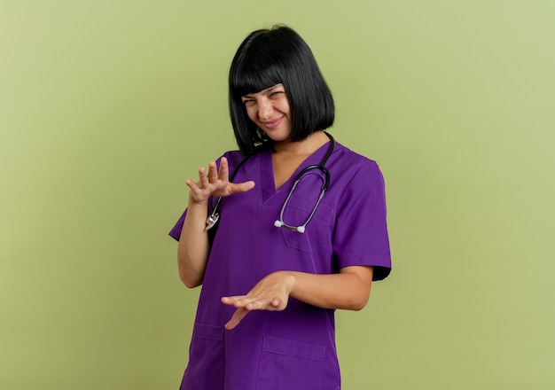 Il giovane medico femminile castana dispiaciuto in uniforme con lo stetoscopio tiene le mani diritte isolate su fondo verde oliva con lo spazio della copia