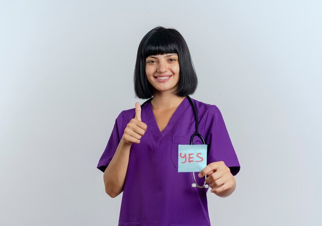 Il giovane medico femminile castana sorridente in uniforme con lo stetoscopio sfoglia in su e tiene la nota di sì