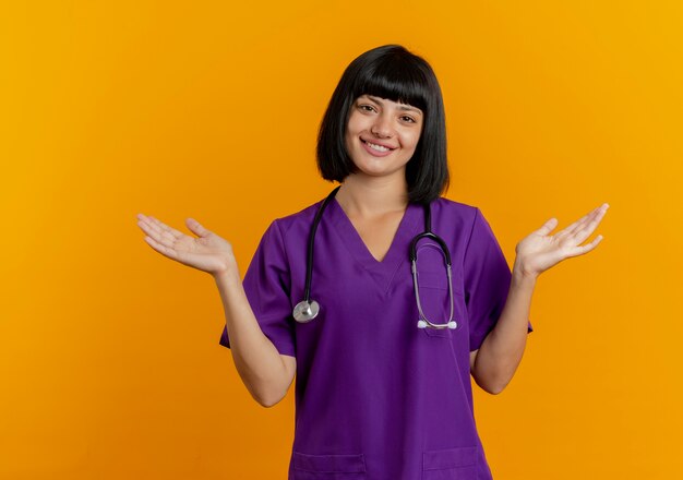 Il giovane medico femminile castana sorridente in uniforme con lo stetoscopio sta con le mani aperte isolate su fondo arancio con lo spazio della copia