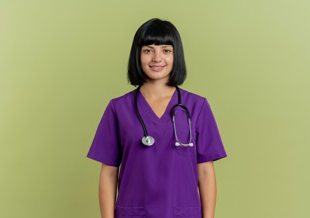 Il giovane medico femminile castana sorridente in uniforme con lo stetoscopio sta isolato su fondo verde oliva con lo spazio della copia