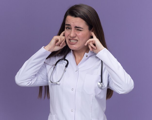 Il giovane medico femminile dispiaciuto che indossa la veste medica con lo stetoscopio chiude le orecchie con le dita sulla porpora