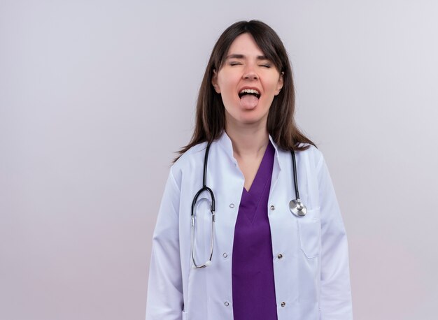 Il giovane medico femminile infastidito in abito medico con lo stetoscopio con gli occhi chiusi sporge la lingua su fondo bianco isolato con lo spazio della copia