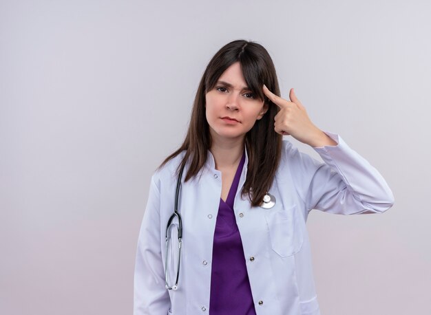 Il giovane medico femminile infastidito in abito medico con lo stetoscopio mette il dito alla tempia su fondo bianco isolato con lo spazio della copia