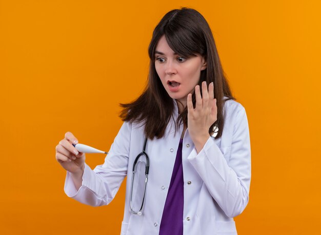 Il giovane medico femminile scioccato in veste medica con lo stetoscopio tiene il termometro su fondo arancio isolato con lo spazio della copia