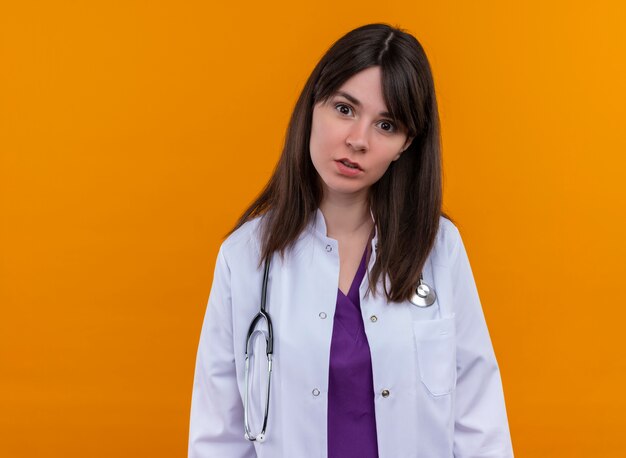 Il giovane medico femminile sorpreso in abito medico con lo stetoscopio esamina la macchina fotografica su fondo arancio isolato con lo spazio della copia