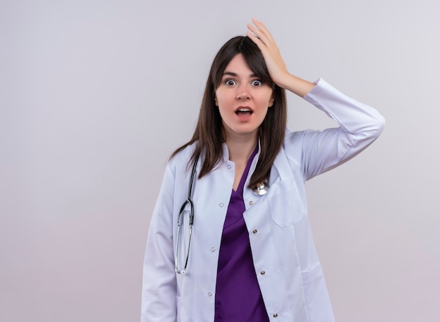 Il giovane medico femminile sorpreso in abito medico con lo stetoscopio mette la mano sulla testa su fondo bianco isolato con lo spazio della copia