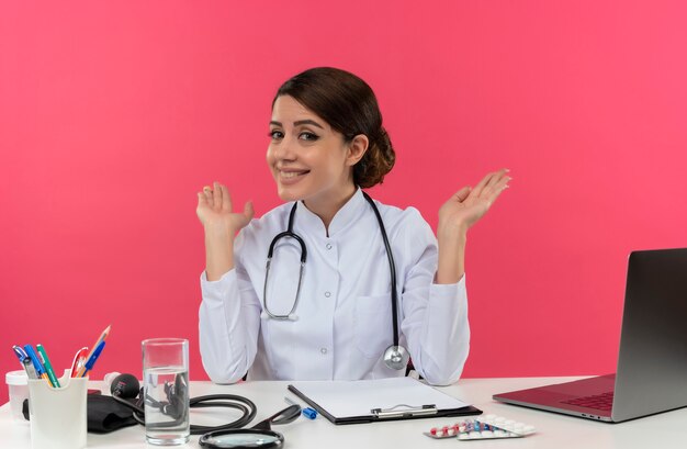 Il giovane medico femminile sorridente che porta veste medica con lo stetoscopio che si siede allo scrittorio funziona sul calcolatore con gli strumenti medici si diffonde le mani con lo spazio della copia