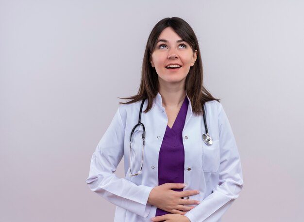 Il giovane medico femminile sorridente in abito medico con lo stetoscopio tiene alla pancia con entrambe le mani su fondo bianco isolato con lo spazio della copia