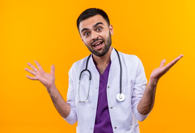 Il giovane medico maschio allegro che indossa l'abito medico dello stetoscopio diffonde le mani sulla parete gialla isolata